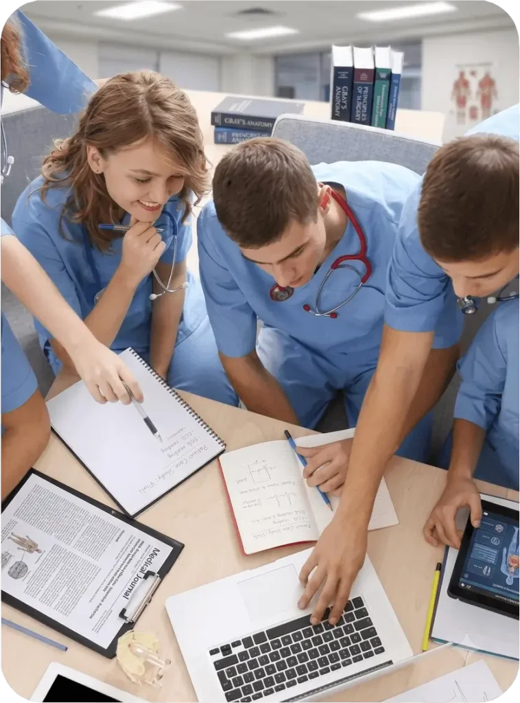 Group of nursing students and professionals wearing blue scrubs collaborating around a table, reviewing medical journals, writing in notebooks, and using a laptop.